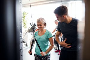 A female senior with a young trainer doing strength workout exercise in gym.